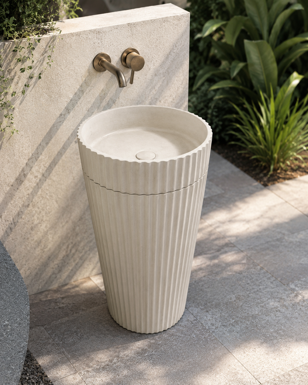 Outdoor fluted concrete pedestal sink with wall-mounted brass faucet on a garden patio.
